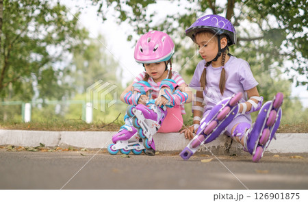 Cute little girls putting on roller skates sitting on the sidewalk. Active sports during the summer holidays 126901875