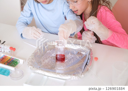 The girls experiment with a test tube and liquids. Great interest. Schoolchildren in protective glasses, huddled close to the table, observe the chemical reaction taking place in the flask. The girls experiment with a test tube and liquids. Great interest. Schoolchildren in protective glasses, huddled close to the table, observe the chemical reaction taking place in the flask. 126901884