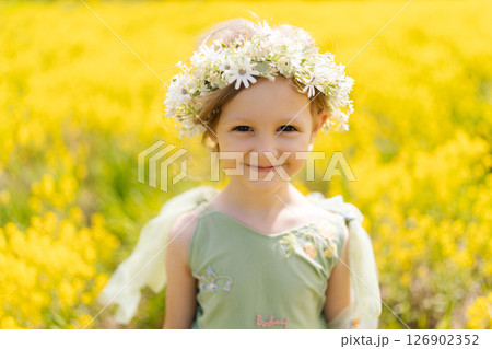 Cheerful preschooler girl wearing beautiful flower crown and fairy dress playing in blooming yellow meadow on sunny summer day, enjoying warm weather and vibrant colors of nature, looking at camera 126902352