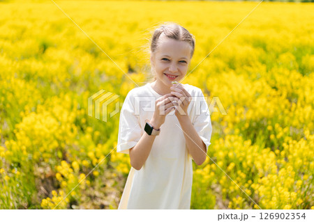 Cheerful young girl 10 yo in white dress, joyfully smelling vibrant yellow flowers standing in warm sunshine of summer day, embracing scents of nature. Concept of happy childhood, outdoor recreation. 126902354