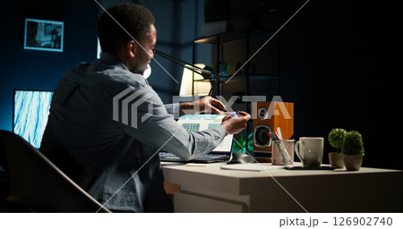 Black man organizing his schedule with a planner and putting a reminder, creating an adhesive note post it and browsing the web. Modern productive guy writes a memo for research. Camera A. 126902740