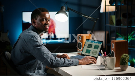 Black accountant working on monthly revenue balance on laptop at home, calculating investment money to double the profit. Economist looks into legal trade statistics, supply and demand. Camera B. 126903364