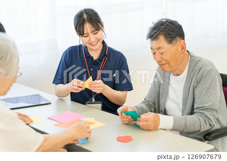 老人ホーム,介護施設で働く介護スタッフの女性と折り紙をするシニア男女 126907679