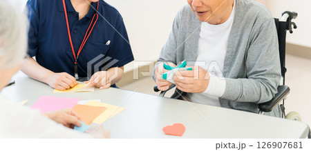 老人ホーム,介護施設で働く介護スタッフの女性と折り紙をするシニア男女 126907681