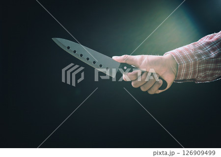 Close-up of male hand in plaid shirt gripping large sharp kitchen knife horizontally in dim lighting on black background Close-up of male hand in plaid shirt gripping large sharp kitchen knife horizontally in dim lighting on black background 126909499