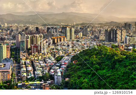 Seen from the lush crest of Xiangshan Mountain, Taipei, Taiwan, the metropolis fans out across its basin in tight rows of low rooftops and clusters of modern high-rise towers, while the forested slope 126910154