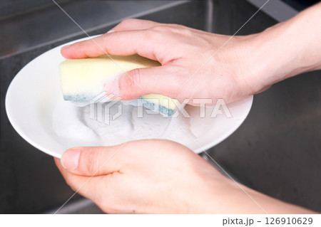 The young man's hands were cleaning dishes and bowls in the sink, and he used a sponge and foam to clean dirt and grease from food. The young man's hands were cleaning dishes and bowls in the sink, and he used a sponge and foam to clean dirt and grease from food. 126910629