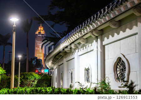 In Taipei, Taiwan, an illuminated skyscraper rises over the white tiled wall of Chiang Kai Shek Memorial Hall, where ornate roof tiles and lattice windows meet palm trees, hedges, and modern city In Taipei, Taiwan, an illuminated skyscraper rises over the white tiled wall of Chiang Kai Shek Memorial Hall, where ornate roof tiles and lattice windows meet palm trees, hedges, and modern city 126911553