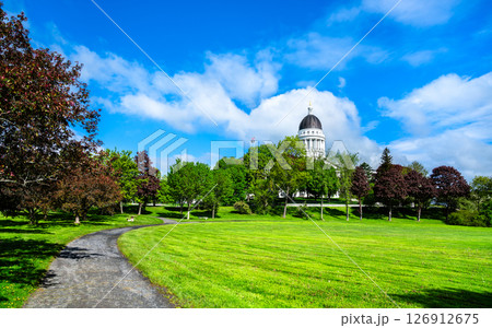 In Augusta, United States, the Maine State House dome peeks above leafy Capitol Park, where a curved gravel path winds across bright spring lawns under a vivid blue sky. 126912675