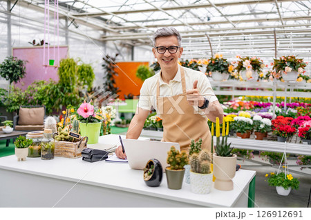 A Caucasian man works at a flower shop's counter showing thumb up, surrounded by a vibrant display of plants and flowers. A Caucasian man works at a flower shop's counter showing thumb up, surrounded by a vibrant display of plants and flowers. 126912691