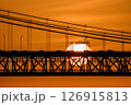 Glowing sun setting behind 25 de Abril bridge, Lisbon, Portugal, suspension cables, cars and bridge structure in silhouette. Bright orange sky, dramatic urban sunset over Tagus River. Close view 126915813