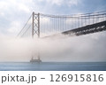 Iconic 25 de Abril suspension bridge, Lisbon, Portugal, in morning fog on sunny day over calm Tagus river waters. Misty atmosphere, peaceful, quiet scene, mist roll in over waters, riverscape, beauty 126915816