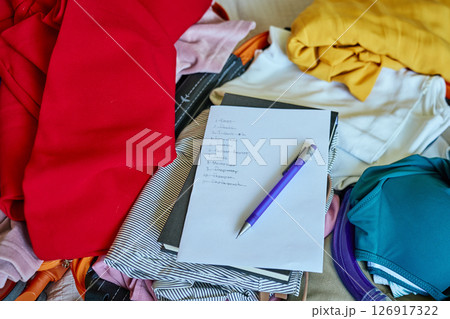 Handwritten checklist and pen on top of colorful folded clothes inside open suitcase. Concept of travel planning and packing essentials for trip 126917322