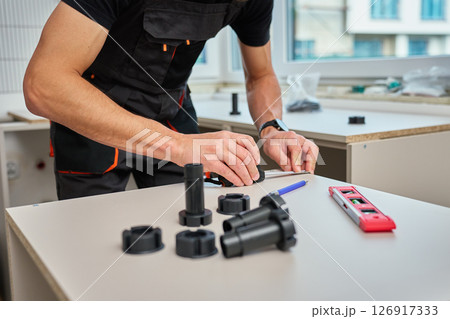 Man measuring furniture part during assembly kitchen cabinets, using measuring tape, tools and plastic furniture legs Man measuring furniture part during assembly kitchen cabinets, using measuring tape, tools and plastic furniture legs 126917333
