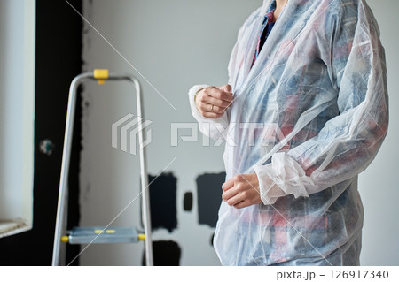 Close-up of woman in protective coveralls preparing to paint wall with color samples in new apartment. Concept of home improvement, renovation and interior updates Close-up of woman in protective coveralls preparing to paint wall with color samples in new apartment. Concept of home improvement, renovation and interior updates 126917340