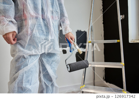 Woman wearing protective coveralls holds black paint roller near painted wall samples and ladder in room under renovation. Painter preparing to paint wall. Home improvement and interior update concept Woman wearing protective coveralls holds black paint roller near painted wall samples and ladder in room under renovation. Painter preparing to paint wall. Home improvement and interior update concept 126917342