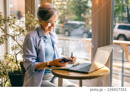 Senior businesswoman writing notes from phone at cafe table with laptop, smiling, working remotely 126917381