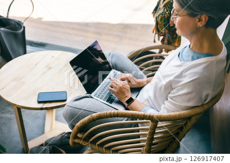 Middle aged woman working on laptop in cozy rattan chair by window with natural light Middle aged woman working on laptop in cozy rattan chair by window with natural light 126917407