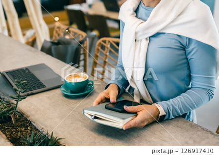 Close-up of woman holding smartphone and notebook in cafe with laptop and coffee, freelance work 126917410