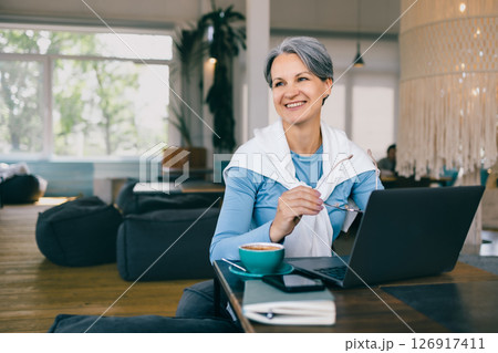 Happy mature woman with gray hair living fully during menopause, working remotely in cozy cafe 126917411