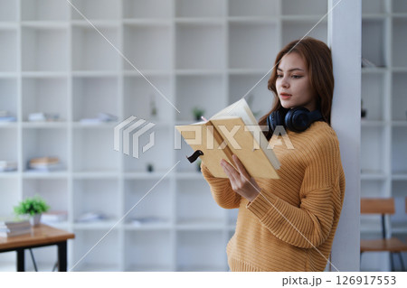 Young woman reads a book with headphones around her neck in a bright room. Young woman reads a book with headphones around her neck in a bright room. 126917553