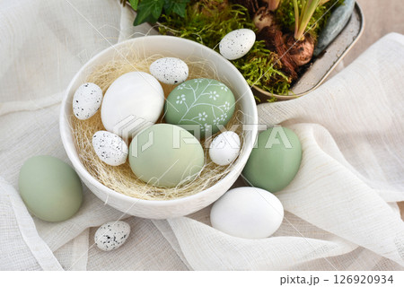 Stylish decorated easter eggs in a white bowl on linen cloth on a light wooden table.  126920934