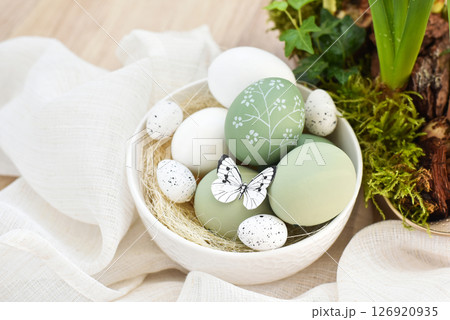 Stylish decorated easter eggs in a white bowl on linen cloth on a light wooden table.  126920935