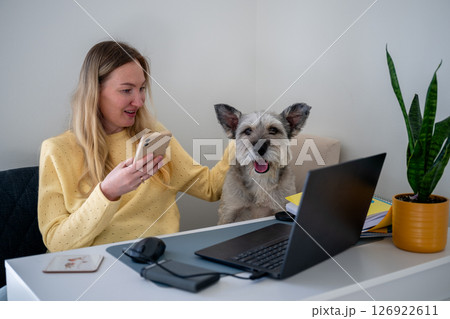 Woman in yellow sweater using phone with dog beside her at home office desk, concept of remote work  126922611