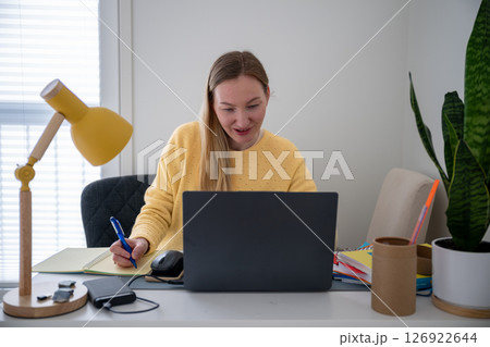 Woman in yellow sweater works on laptop and writes notes at a home office desk with lamp and plant  126922644