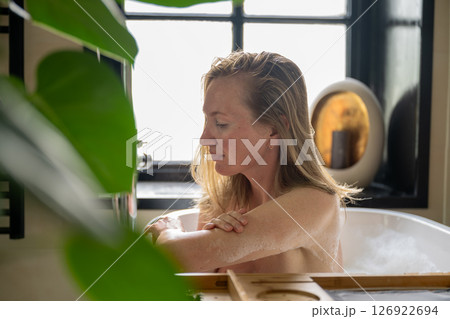 Woman relaxing in bathtub near window with plants and candles around her 126922694