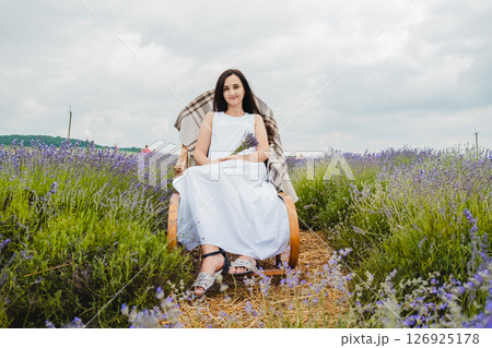 A young woman in a long white dress sits on a rocking chair in the middle of a lavender field 126925178