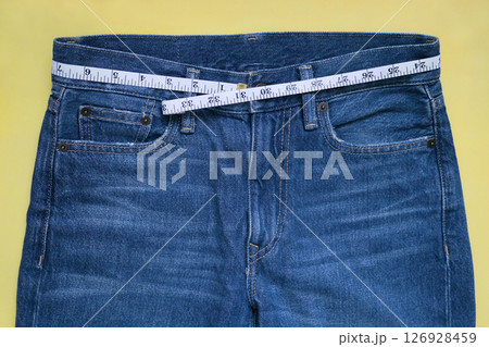 Female blue jeans with measuring tape flat lay on a yellow background. Losing weight, overweight and healthy lifestyle concept Female blue jeans with measuring tape flat lay on a yellow background. Losing weight, overweight and healthy lifestyle concept 126928459