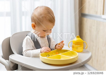 Sweet little baby boy sitting on his chair and eating something from the plate Sweet little baby boy sitting on his chair and eating something from the plate 126928846