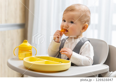 Sweet little baby boy sitting on his chair and eating something from the plate Sweet little baby boy sitting on his chair and eating something from the plate 126928847