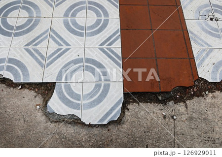 a close-up view of a pavement featuring a white and blue round and diamond shape pattern tile and a brown tiles. The edges of the tiles are chipped and uneven, revealing the underlying surface. a close-up view of a pavement featuring a white and blue round and diamond shape pattern tile and a brown tiles. The edges of the tiles are chipped and uneven, revealing the underlying surface. 126929011