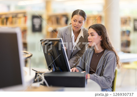 Female student works on computer in library and talks with female friend. 126930737