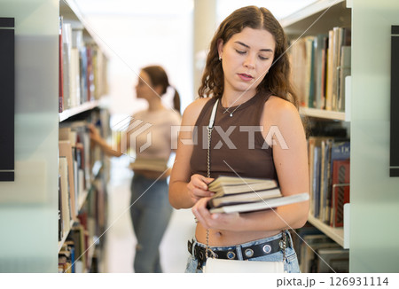 Young female student stands in library with stack of books Young female student stands in library with stack of books 126931114