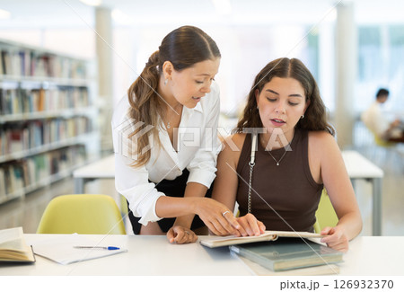Two female student sits at table in library and studies, reads books and takes notes Two female student sits at table in library and studies, reads books and takes notes 126932370