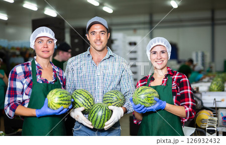Smiling factory workers holding ripe whole watermelons and looking at camera during sorting ripe watermelons on conveyor Smiling factory workers holding ripe whole watermelons and looking at camera during sorting ripe watermelons on conveyor 126932432