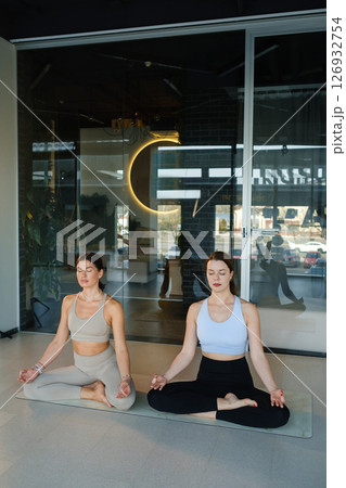 Two beautiful girls doing yoga in lotus pose. Two girls meditating at yoga sitting in lotus pose in the fitness center Two beautiful girls doing yoga in lotus pose. Two girls meditating at yoga sitting in lotus pose in the fitness center 126932754