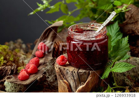 A jar of raspberry jam with fresh berries on an old tree trunk in the forest. A jar of raspberry jam with fresh berries on an old tree trunk in the forest. 126933918