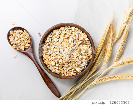 Top view of a wooden bowl with rolled oats and a wooden spoon, dried wheat stalks on a light marble background. Top view of a wooden bowl with rolled oats and a wooden spoon, dried wheat stalks on a light marble background. 126935505