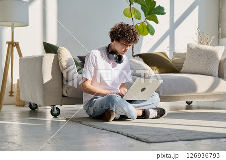 Handsome young male typing on laptop sitting on floor at home Handsome young male typing on laptop sitting on floor at home 126936793