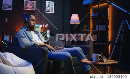 Black teleworker putting headset to listen to webinar discussion, participates at an online lesson on laptop to improve his skills. Young person enjoying meeting from his living room. Camera A. 126938109