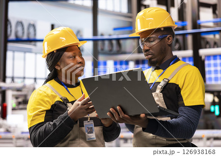 In smart manufacturing plant, african american engineers supervise industrial automation system using laptop. The automated factory features conveyor belts, robotic arms and optimized assembly lines. 126938168