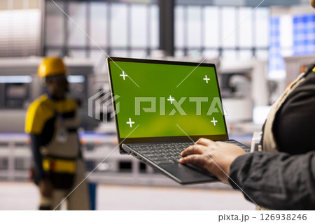 African american factory worker holds laptop with green screen display, overseeing automated production line. The industrial plant integrates robotics, conveyor belts and smart manufacturing systems. 126938246