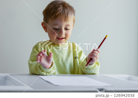 Cheerful little boy in a yellow sweater draws with colored pencils on paper at a table 126942038