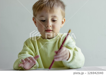 Cheerful little boy in a yellow sweater draws with colored pencils on paper at a table 126942040