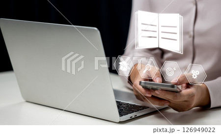 Businesswoman using a cellphone and showing a book icon. Laptop on the table. Technology innovation on a virtual screen. Concept of E-library Businesswoman using a cellphone and showing a book icon. Laptop on the table. Technology innovation on a virtual screen. Concept of E-library 126949022