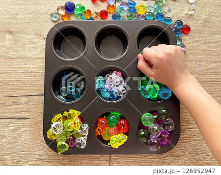 A child's hand arranges glass pebbles in a black metal mold. The child plays while sitting at a wooden table. 126952947